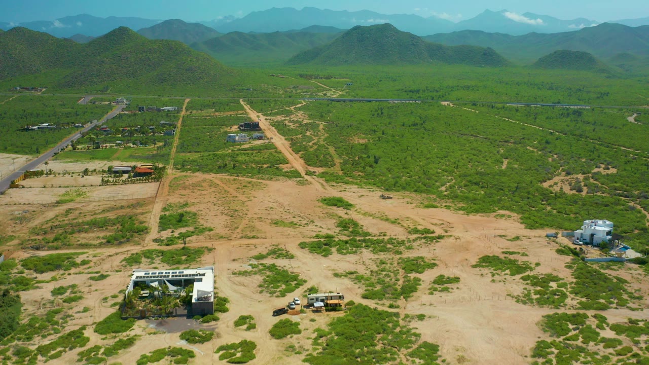 High aerial frame over Cerritos Beach, Baja California Sur showing tan dirt roads through green scrubland, scattered structures, small vehicles, a white building near the lower edge, distant mountains, and no visible people.