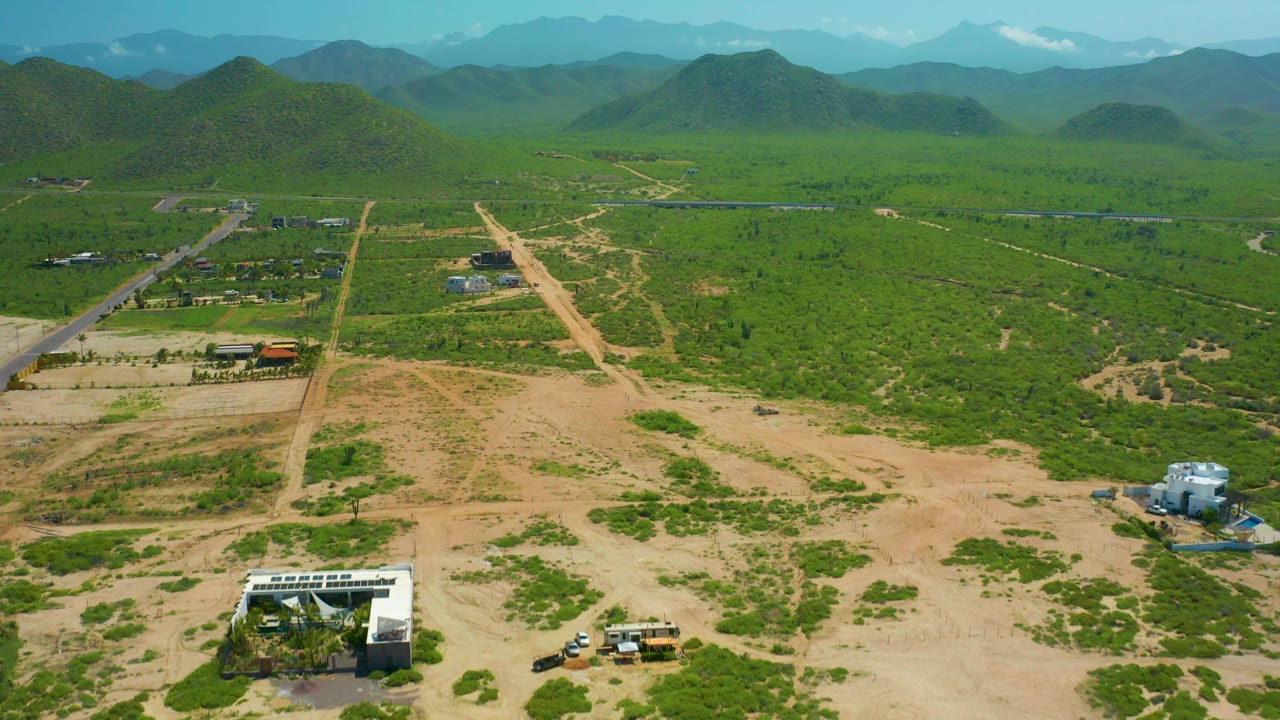 Wide aerial frame in Cerritos Beach, Baja California Sur showing sandy open lots in the foreground, patchy green vegetation, intersecting dirt roads, scattered rural structures, distant mountains, and no visible people.