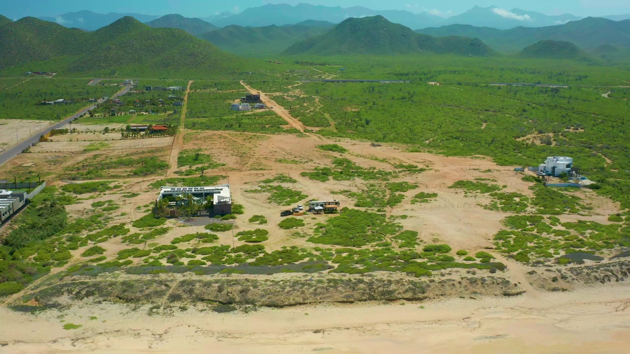 Opening aerial frame over Cerritos Beach, Baja California Sur, showing sandy open terrain, green scrub, dirt roads, scattered buildings, distant hills, and no visible people.
