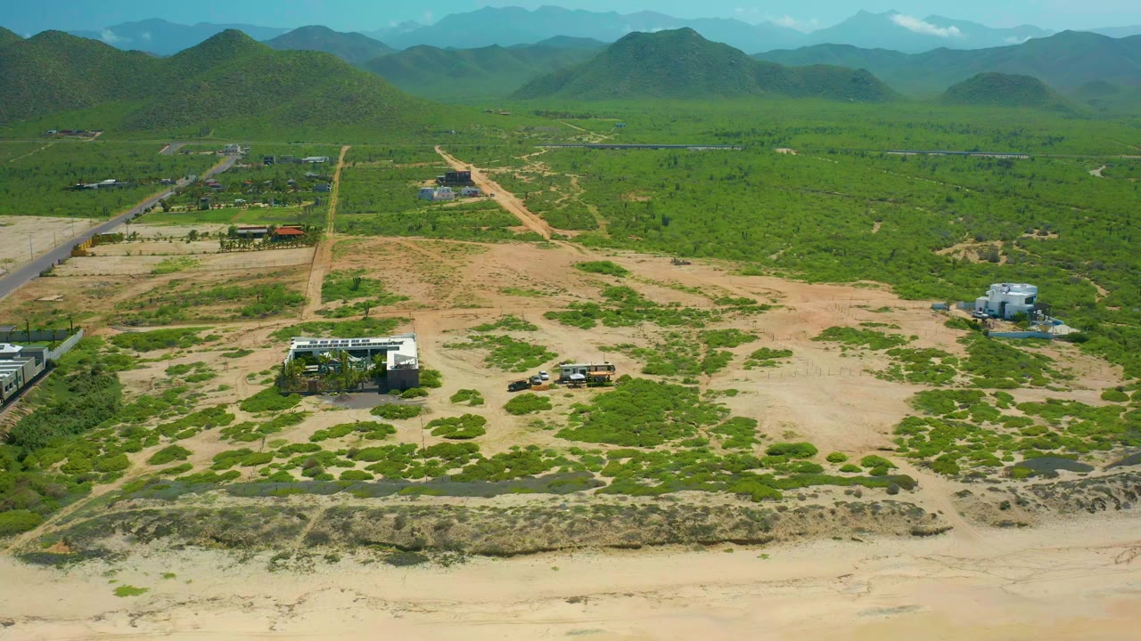 Aerial frame at Cerritos Beach, Baja California Sur, with a dirt road running into green vegetation, sandy lots in the foreground, low buildings at left and right, distant hills, and no visible people.