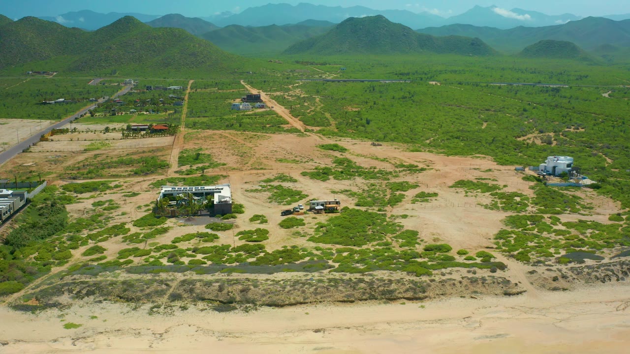 Wide aerial frame in Cerritos Beach, Baja California Sur, showing sandy foreground, scattered green vegetation, pale dirt roads, a few small structures, mountain ridges, and no visible people.