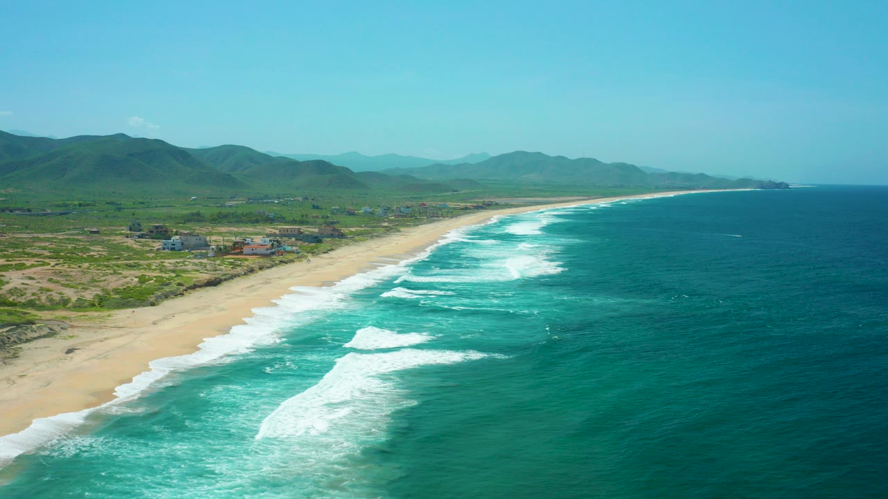 Aerial view of Cerritos Beach in Baja California Sur with turquoise surf rolling diagonally onto sand, green hills inland, scattered buildings near the shore, and no visible people.
