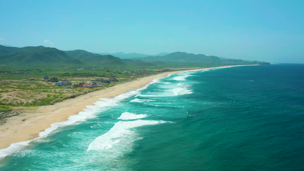 Cerritos Beach aerial frame showing a broad band of turquoise water, white wave breaks closer to shore, tan sand curving into the distance, inland hills, and no visible people.