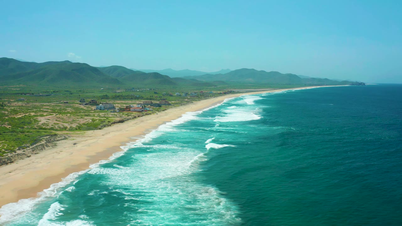High aerial frame over Cerritos Beach showing foamy waves reaching the sand, deep teal water offshore, green scrubland and small structures inland, hazy mountains, and no visible people.