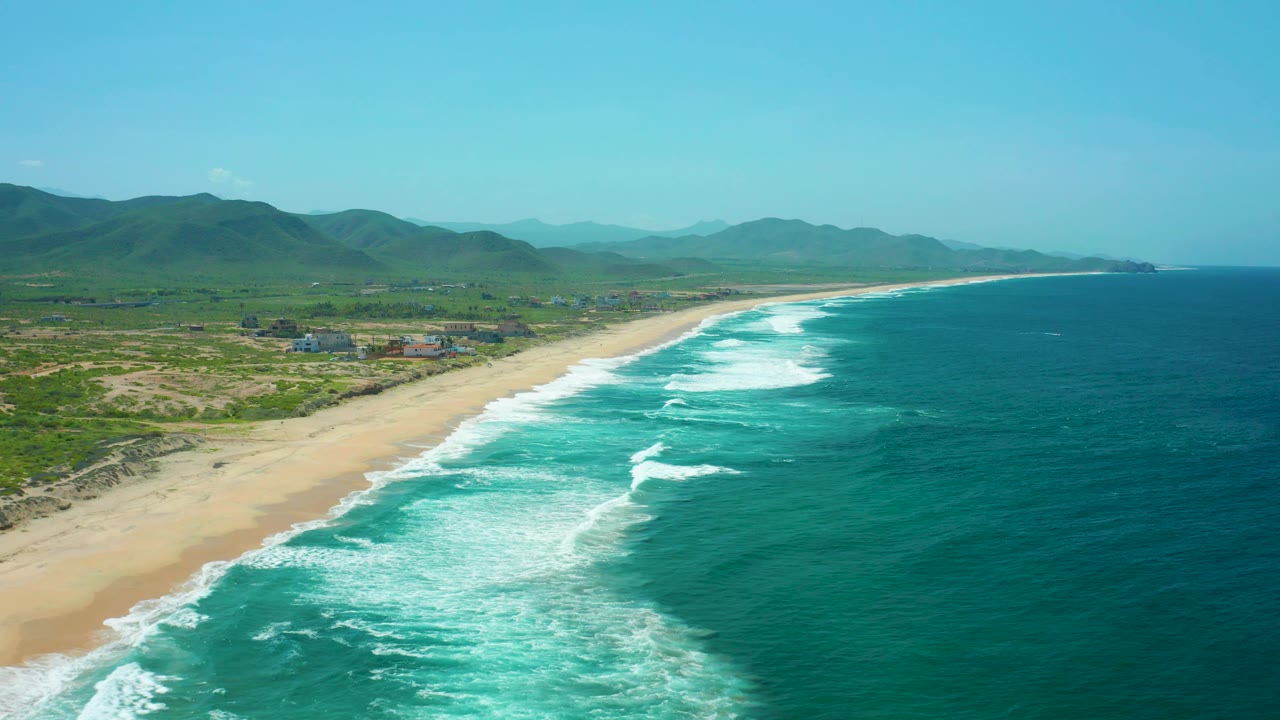 Drone aerial over Cerritos Beach with the sandy shoreline angling into the distance, white wave sets across turquoise water, green hills inland, mountains on the horizon, and no visible people.
