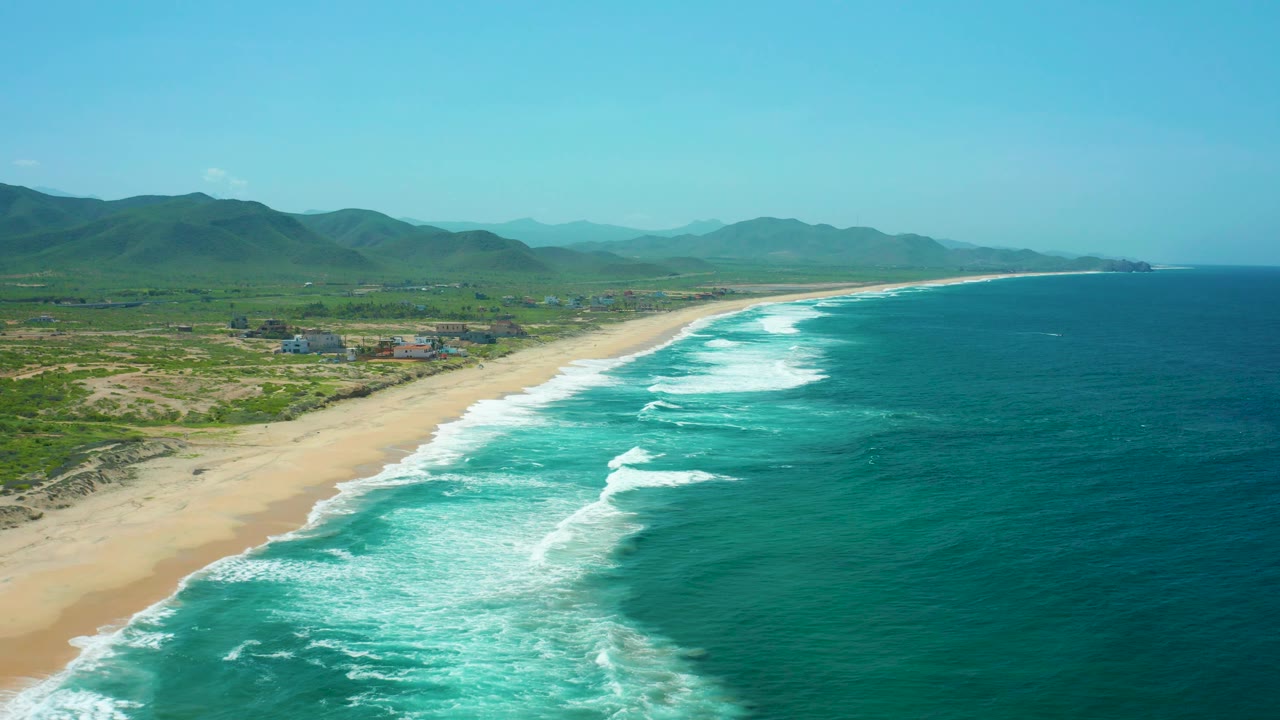 Aerial view of Cerritos Beach with a wide band of white surf near the lower frame, pale sand beside low dunes, green inland terrain, distant mountains, and no visible people.