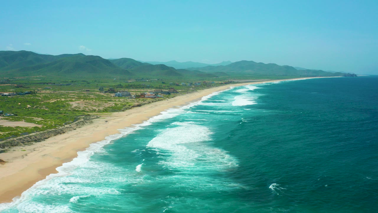 Aerial view over Cerritos Beach in Baja California Sur, with turquoise ocean, white surf bands, sandy shore, green vegetation, distant mountains, and no visible people.