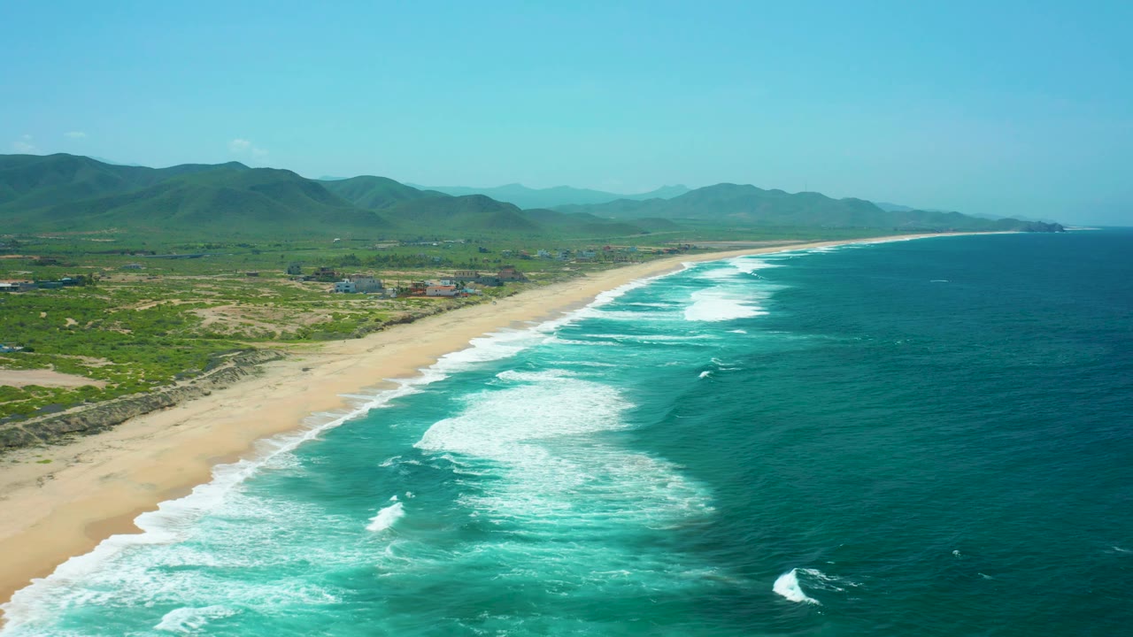 Wide aerial view of Cerritos Beach with darker turquoise water at right, curved white wave lines near shore, pale sand, green coastal land, mountains, and no visible people.