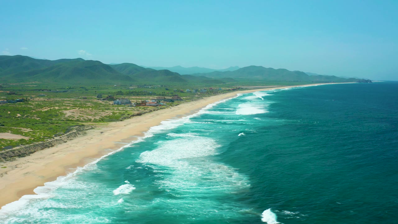 Drone-height view along a sandy coast with foamy surf and green mountains inland.