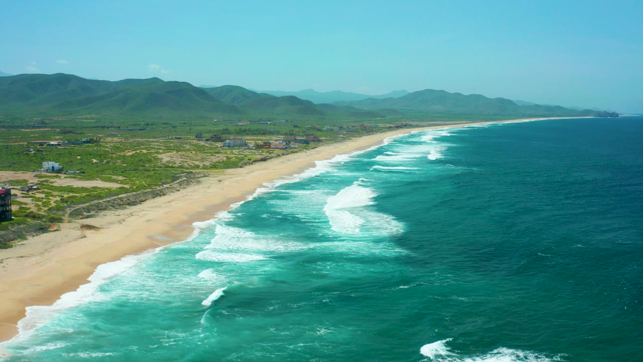 Opening aerial frame over Cerritos Beach, Baja California Sur, with turquoise surf sweeping toward a wide sand shoreline, green inland hills, low buildings, clear sky, and no visible people.