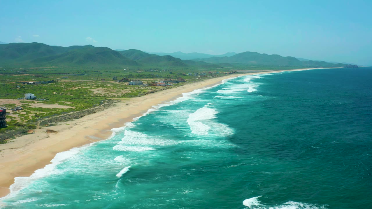 Aerial view one second into the Cerritos Beach clip, showing longer white wave bands along the sand, darker turquoise water offshore, inland green hills, scattered structures, and no visible people.