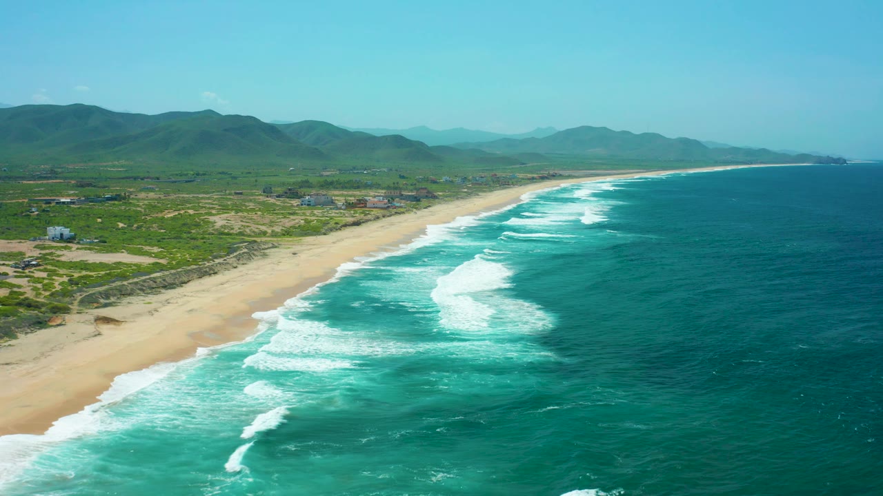 Cerritos Beach appears from a high oblique angle with foamy surf breaking near the sand, broad blue-green ocean to the right, green hills inland, hazy distant mountains, and no visible people.