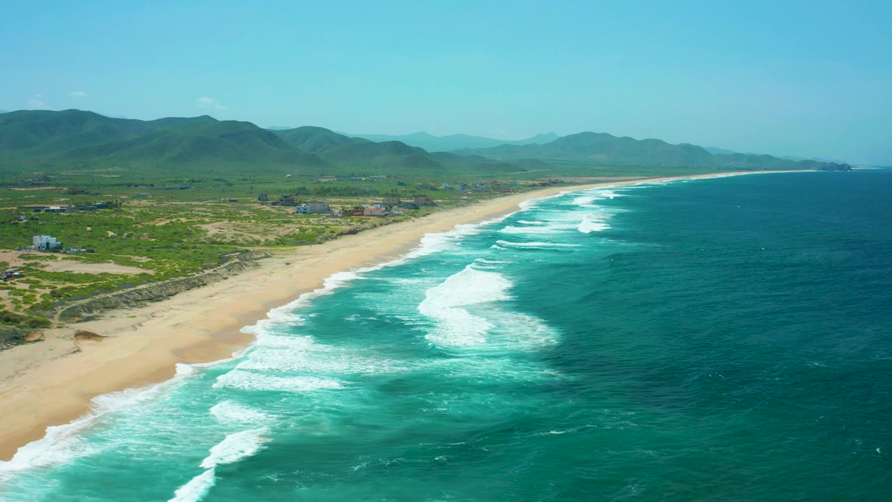 Mid-clip aerial frame of Cerritos Beach with a long sand strip on the left, turquoise breakers curling in from the right, green hills across the background, sparse development inland, and no visible people.