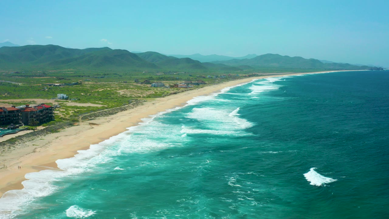 Opening aerial frame over Cerritos Beach, Baja California Sur, with turquoise surf on the right, a long sandy beach on the left, green hills in the distance, and no visible people.