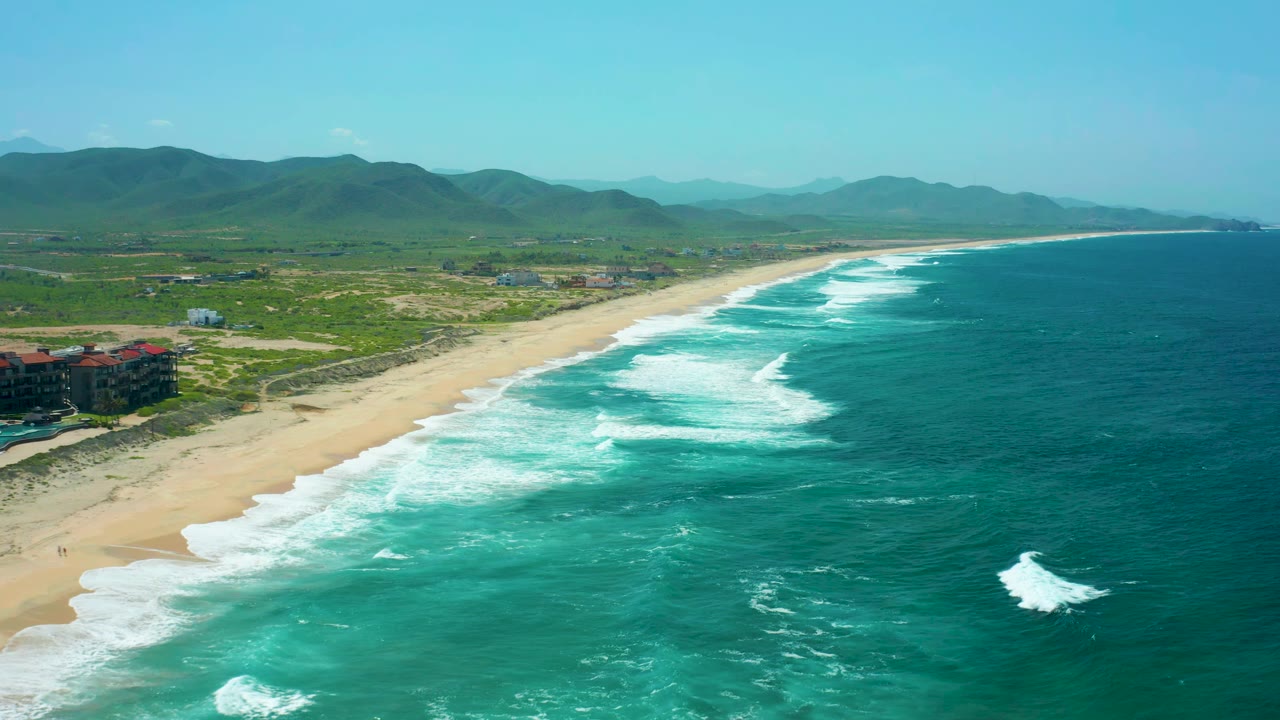 Aerial coastline with white wave bands rolling toward a wide sandy shore.