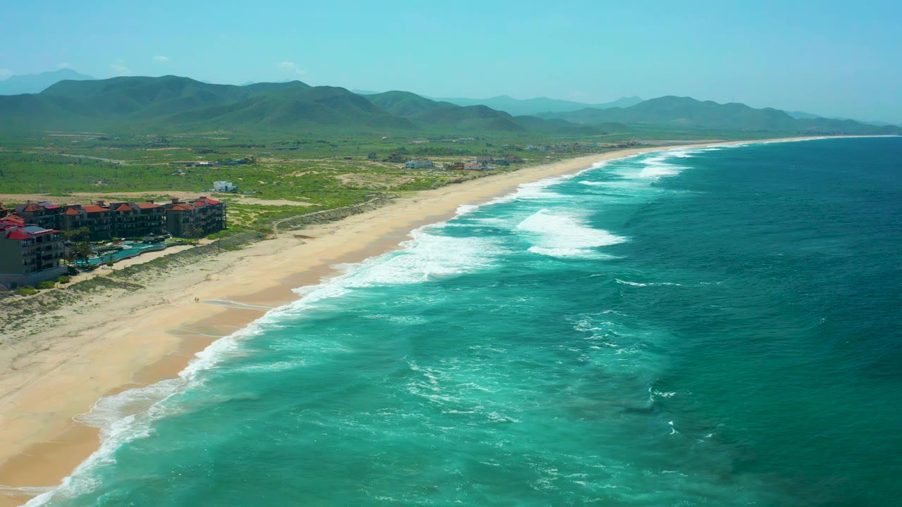 Cerritos Beach appears from above with repeated white wave bands crossing turquoise water, a broad sand strip, low buildings near the dunes at left, hazy hills beyond, and no visible people.