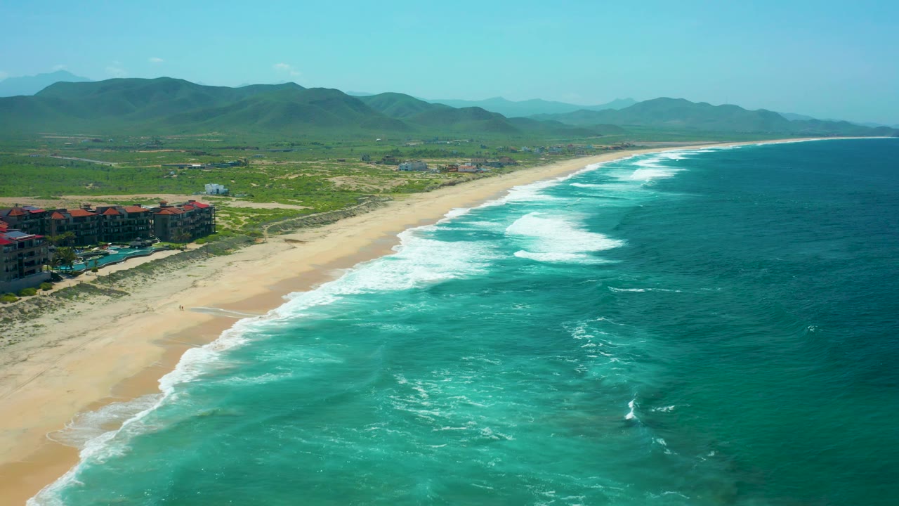 A drone-height view looks down the Cerritos Beach shoreline as white waves break in angled lines, ocean fills the right side, low buildings sit left of the sand, mountains line the horizon, and no visible people appear.