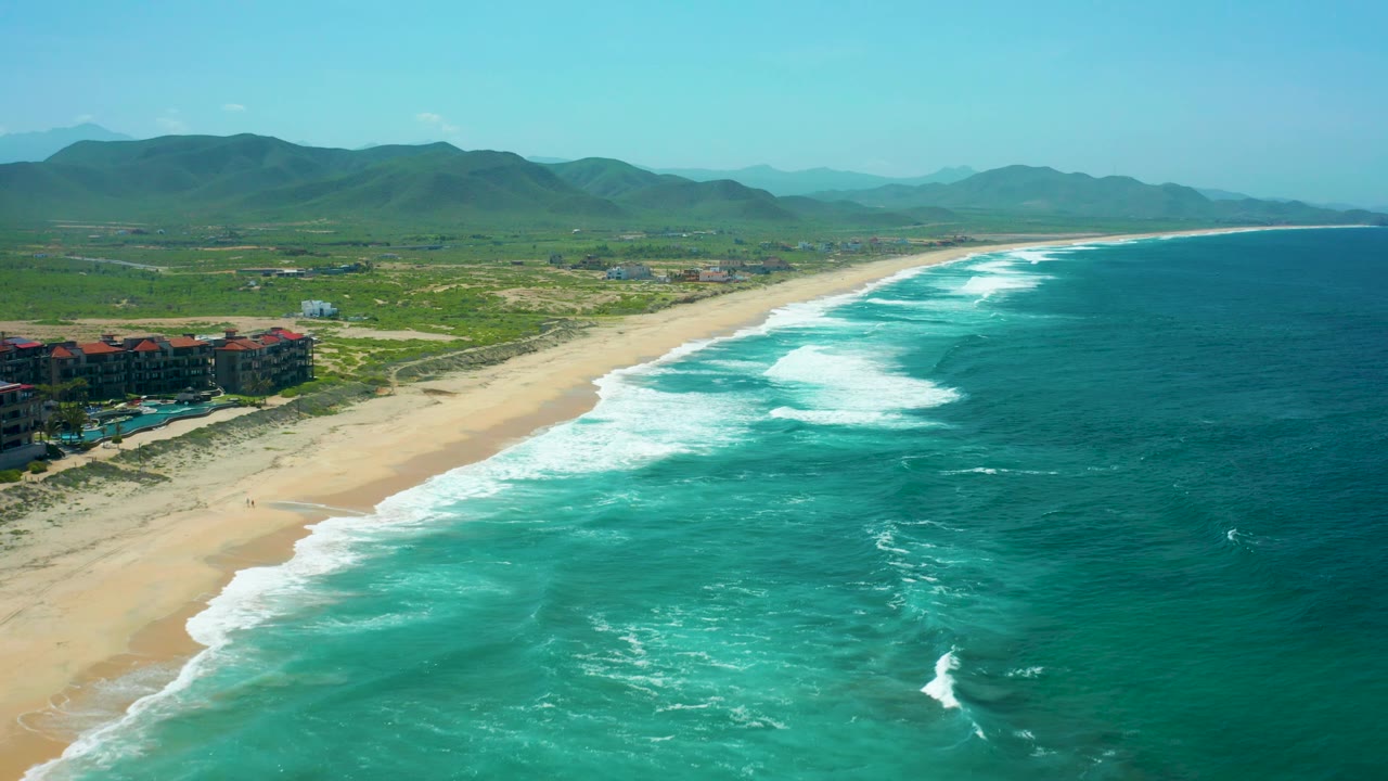 Aerial beach scene with turquoise ocean, white surf, dunes, buildings, and hills.