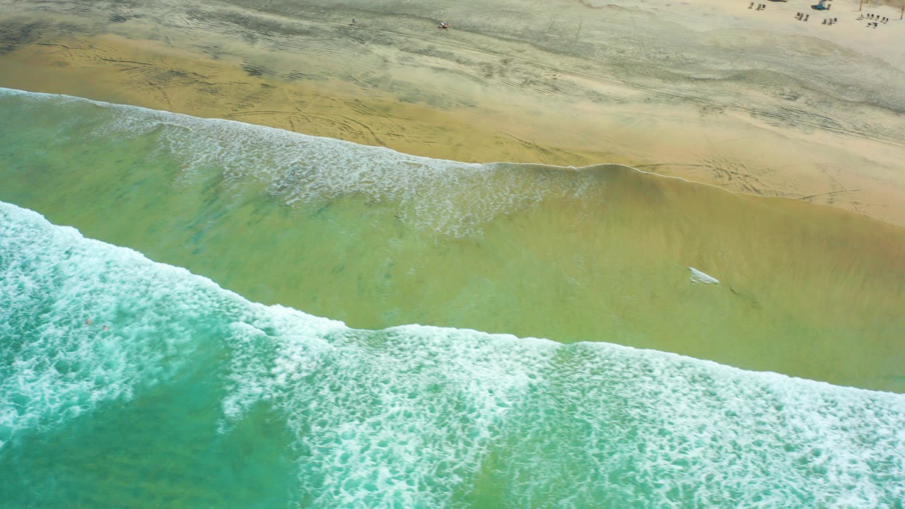 High aerial view at Cerritos Beach, Baja California Sur, Mexico, with turquoise surf, green shallows, tan sand, and a small surfboard or surfer near the center of the break.