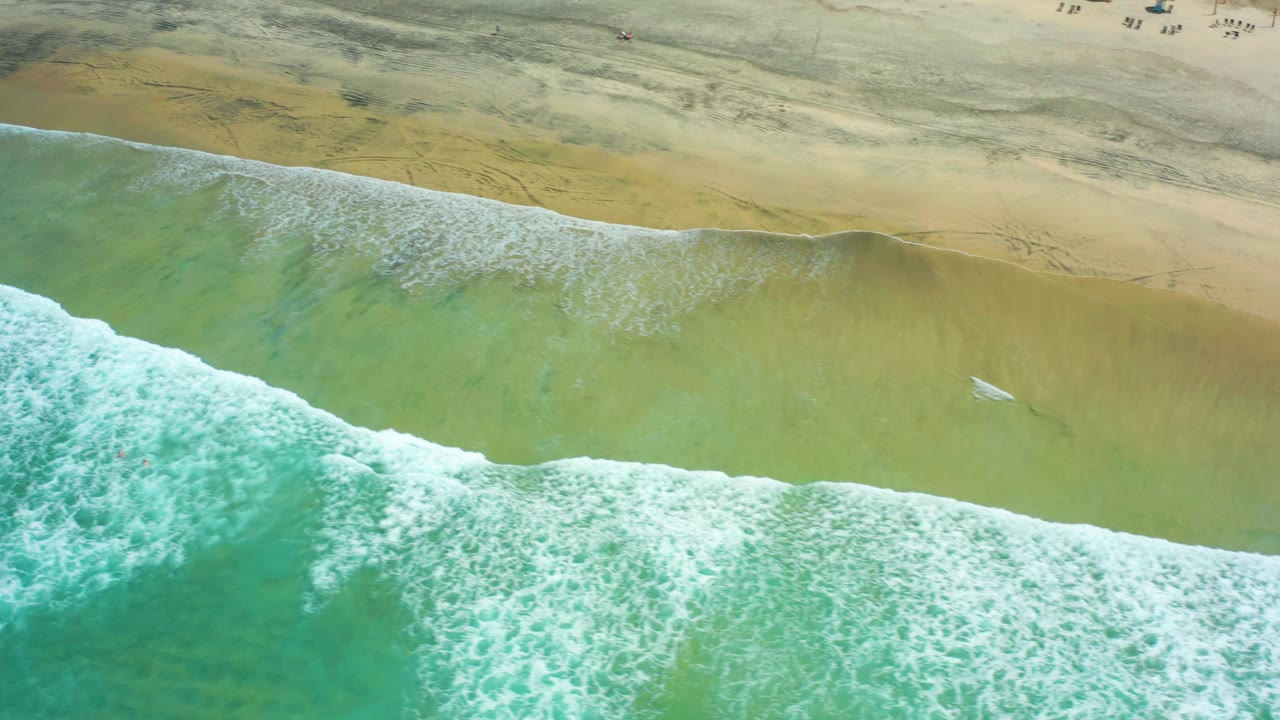 Aerial beach view with a breaking wave and small surfboard in the shallows.