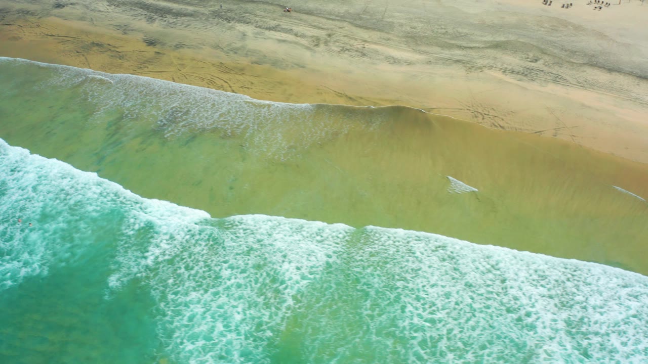 Aerial view of Cerritos Beach as foamy turquoise water fills the lower frame, green shallows sit near the sand, and a small surfboard or surfer appears near the right side of the water.