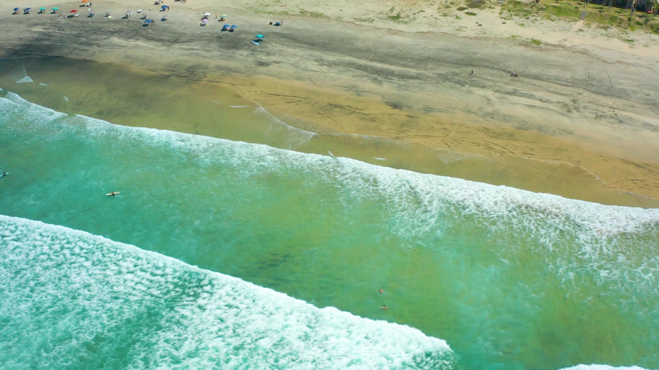 Overhead view of Cerritos Beach with turquoise surf, white foam, wet sand, scattered umbrellas, and small beachgoers visible near the upper edge.