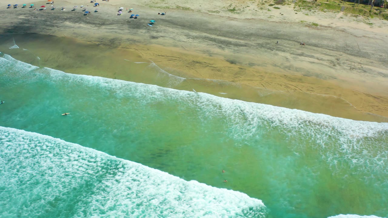 Turquoise wave foam crosses toward the beach with small figures near the sand.