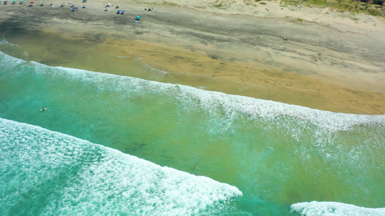 Overhead frame of Cerritos Beach with a diagonal band of white surf crossing green-blue water toward broad sand, with a few distant beachgoers near the top edge.