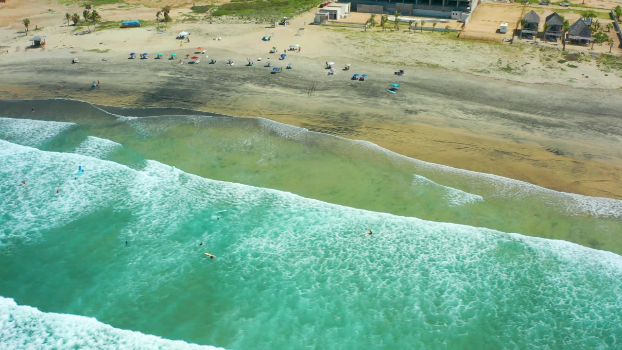 Aerial view over Cerritos Beach in Baja California Sur, with turquoise water and white surf in the foreground, swimmers or surfers offshore, and scattered beach chairs along the sand.