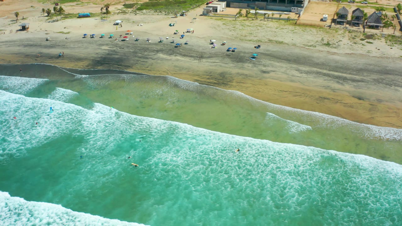 Drone view of foamy waves, shallow green water, and beach chairs on sand.