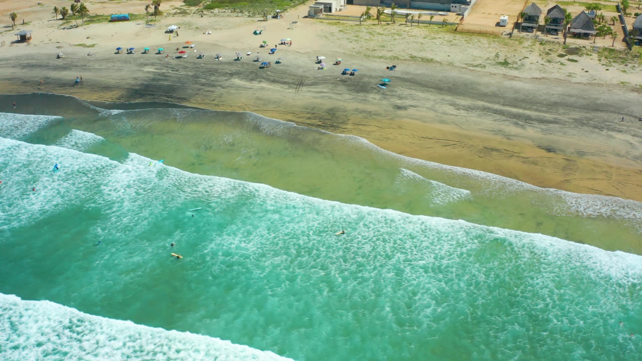 Aerial frame from Cerritos Beach in Baja California Sur, with a diagonal white surf band, turquoise water below it, swimmers or surfers as small figures, and beach chairs and structures along the upper sand.