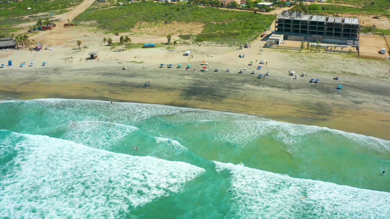 Opening frame over Cerritos Beach, Baja California Sur, with turquoise water, white foam, wet sand, scattered umbrellas, small visible beachgoers, and a large structure behind the shore.