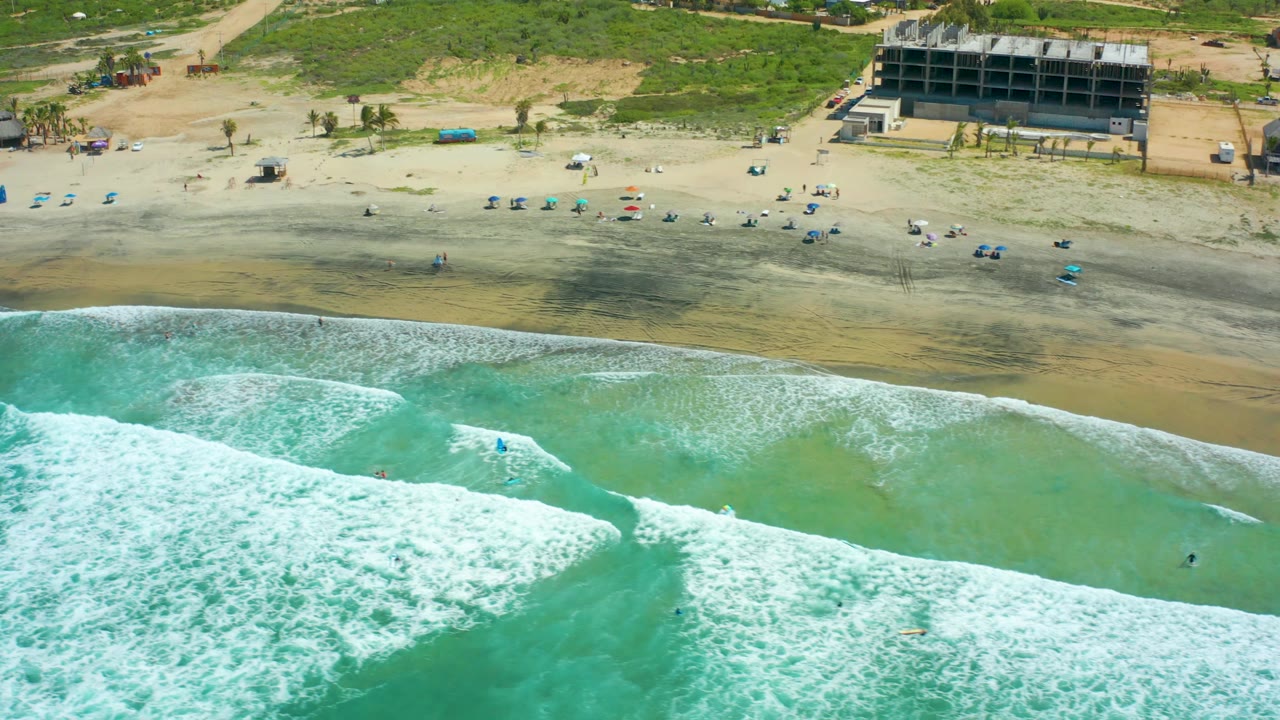 Cerritos Beach, Baja California Sur, seen from above as a thicker white wave band reaches the lower frame, with pale wet sand, scattered blue umbrellas, small people, and vegetation behind the beach.