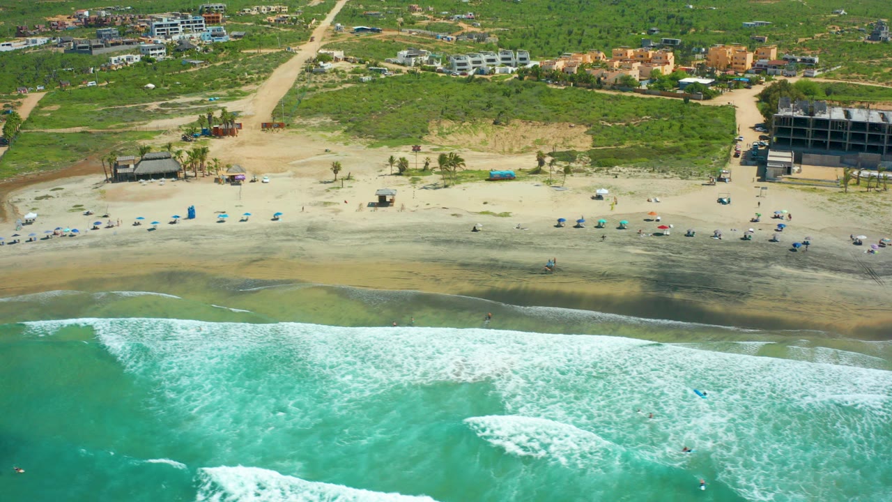 Aerial frame over Cerritos Beach, Baja California Sur, showing turquoise water and white foam in the foreground, a sandy beach with small umbrellas and distant people, and low-rise buildings behind dune greenery.