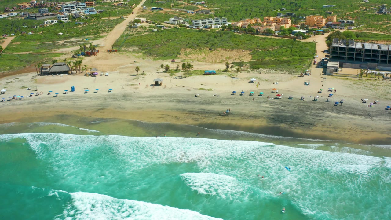 Cerritos Beach appears from above with a broad white wave breaking in turquoise water, small figures near umbrellas on the sand, and a strip of greenery and low buildings beyond the beach.