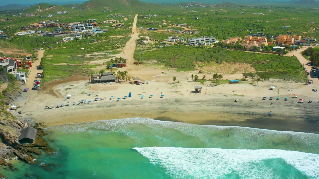 Aerial shoreline view with white surf spreading toward sand and buildings behind the beach.