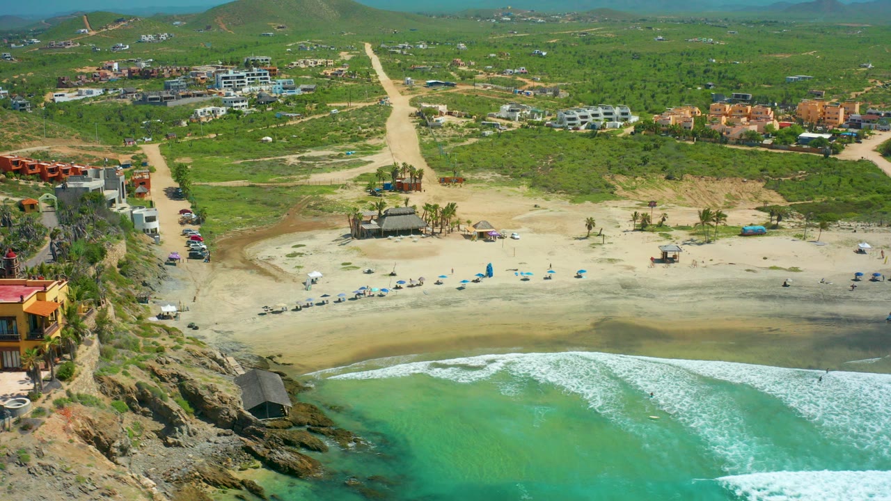 At 0:00, an aerial view over Cerritos Beach shows turquoise surf in the foreground, a sandy beach with chairs and shade structures, inland roads, buildings, green hills, and no visible people.