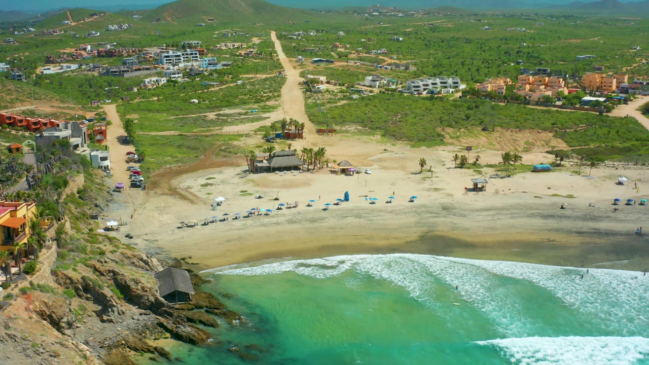 At 0:02, the aerial view of Cerritos Beach shows a broad white wave band crossing the lower frame, pale sand with chairs and shade structures, dirt roads, scattered buildings, green hills, and no visible people.