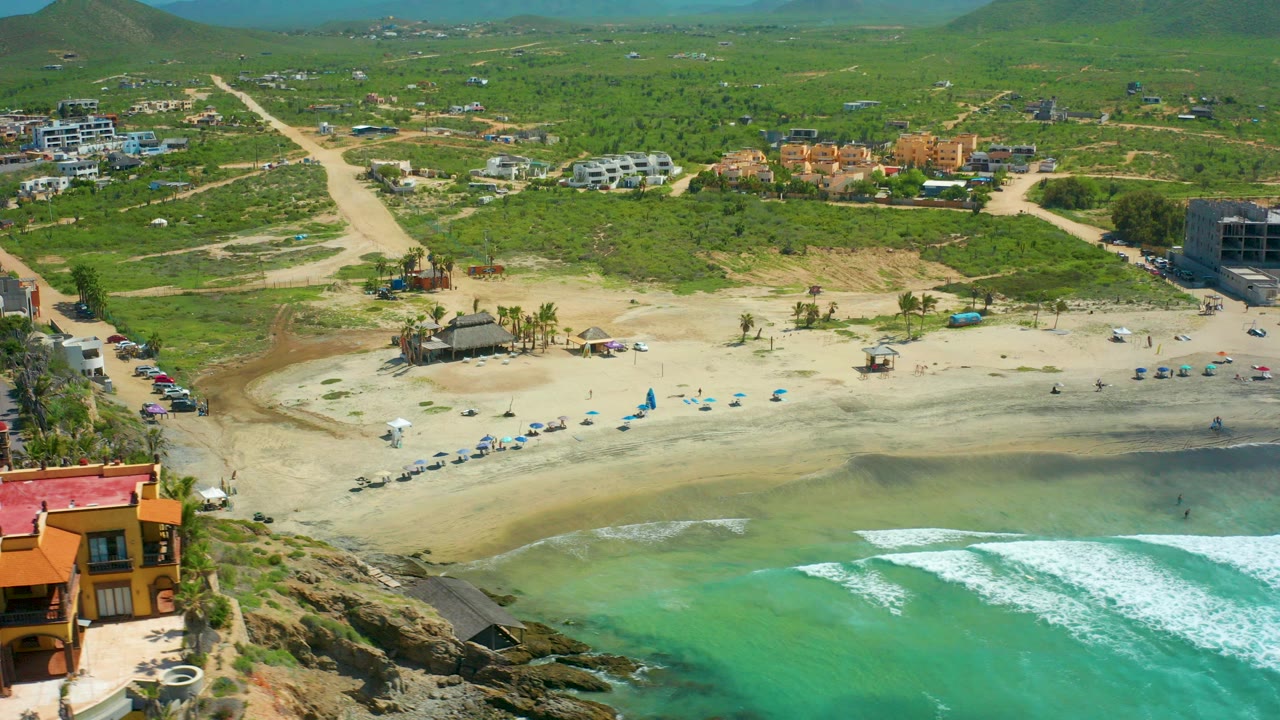 Aerial frame over Cerritos Beach in Baja California Sur showing turquoise surf at the bottom, pale sand with blue umbrellas, road access, scattered buildings, green hills, and no visible people.