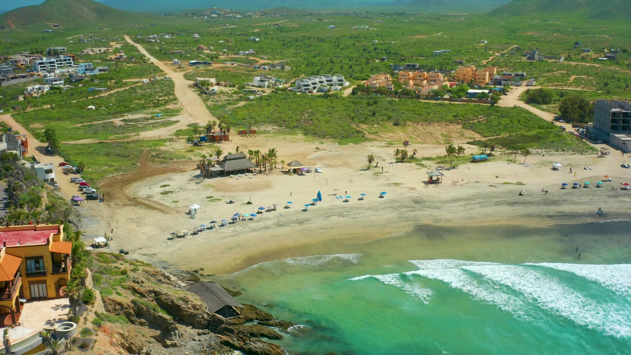 High aerial frame at Cerritos Beach showing turquoise water and white surf along the lower edge, a sandy beach with blue umbrellas, road access, parked cars, low buildings, green hills, and no visible people.