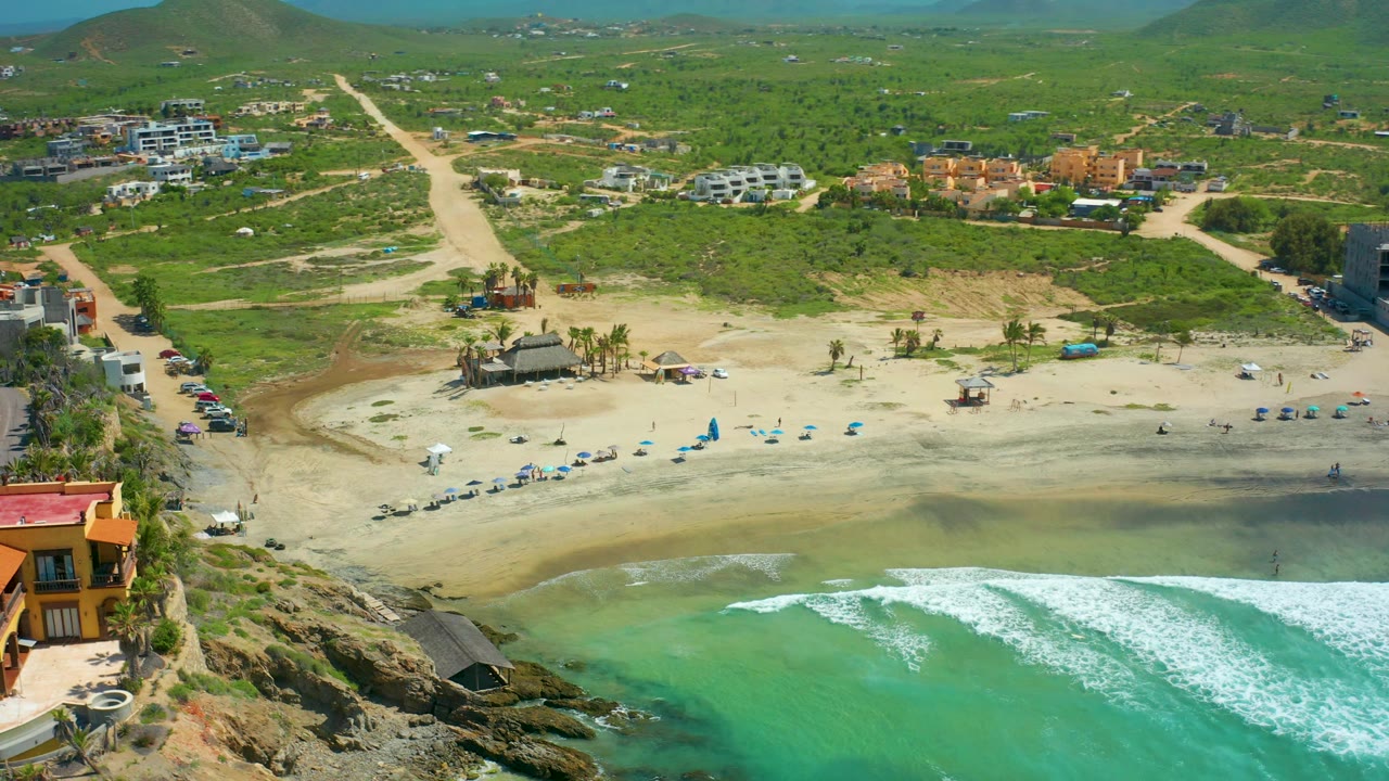 Aerial coastline with wave foam, beach umbrellas, parked cars, buildings, dirt roads, and green hills.