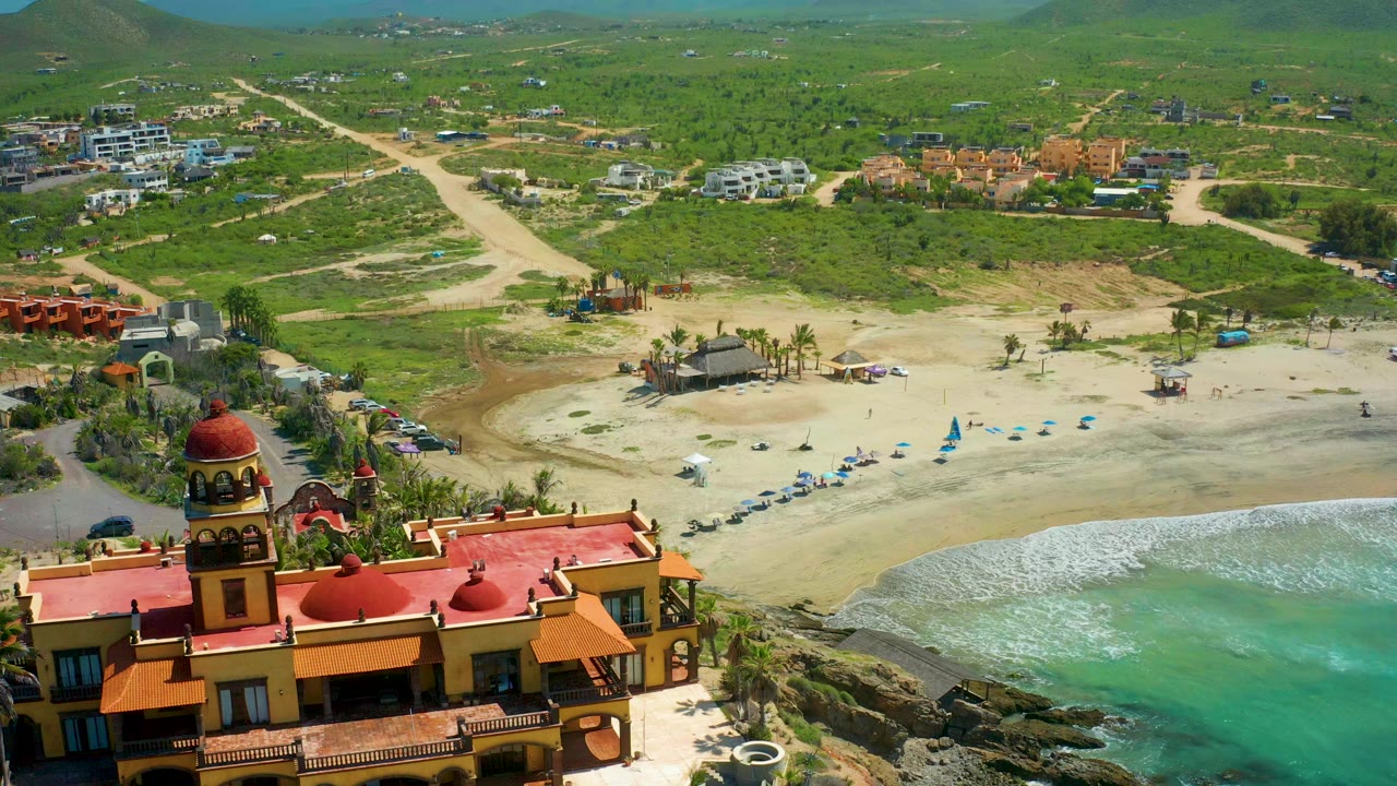 Aerial frame over Cerritos Beach, Baja California Sur, with a red-roofed building in the lower left foreground, blue umbrellas on the sand, turquoise water at right, green hills beyond, and no visible people.