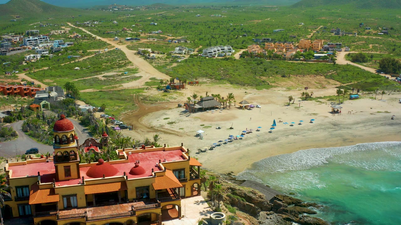 High oblique frame over Cerritos Beach with red roofs along the lower edge, pale sand across the center, small blue umbrellas near the shore, turquoise water at right, and no visible people.