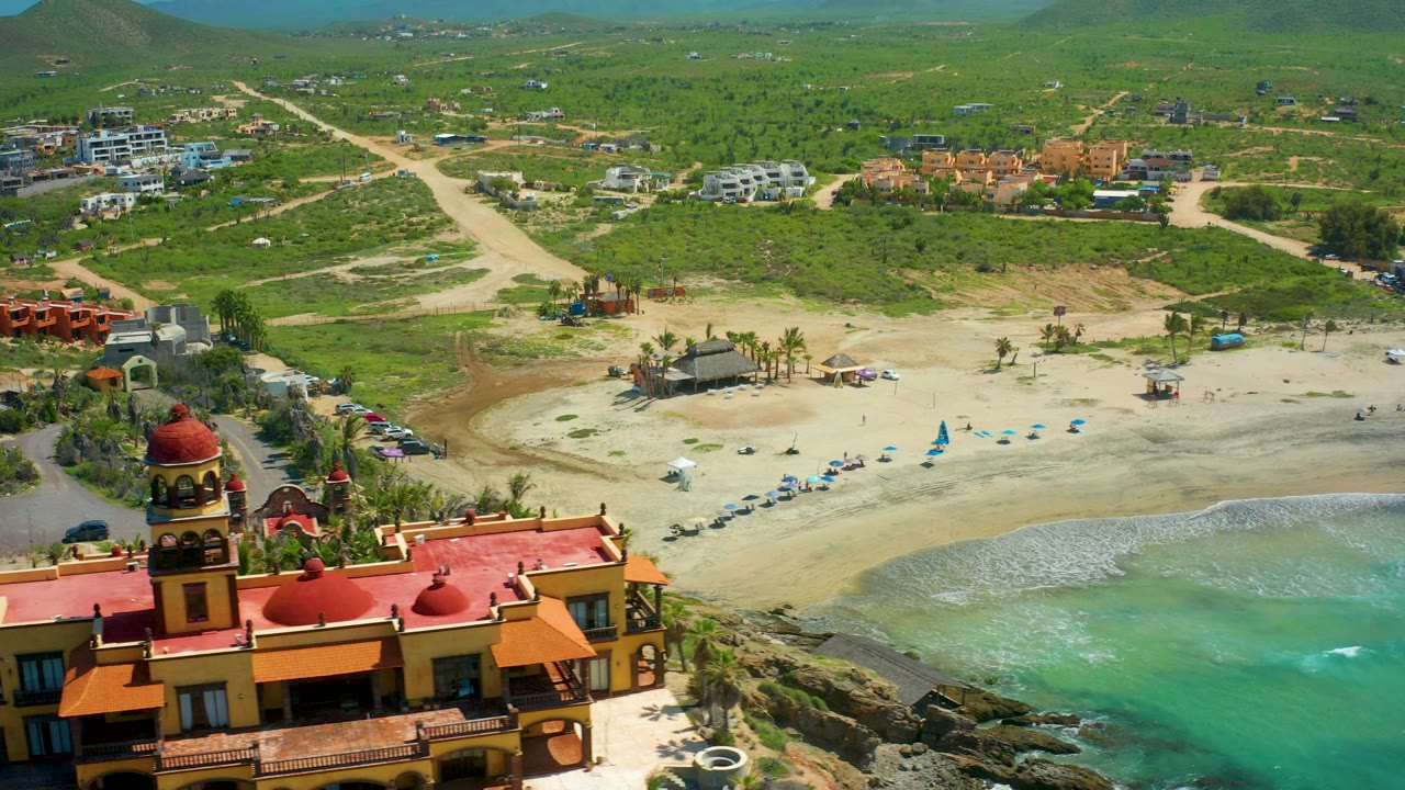 Aerial coastline with red-roofed building lower left, sandy beach, umbrellas, waves, and hills.