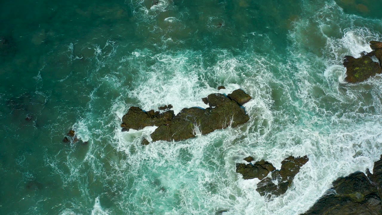 Overhead view at Cerritos Beach, Baja California Sur, showing turquoise water and white surf wrapping around dark rocky outcrops, with no visible people.