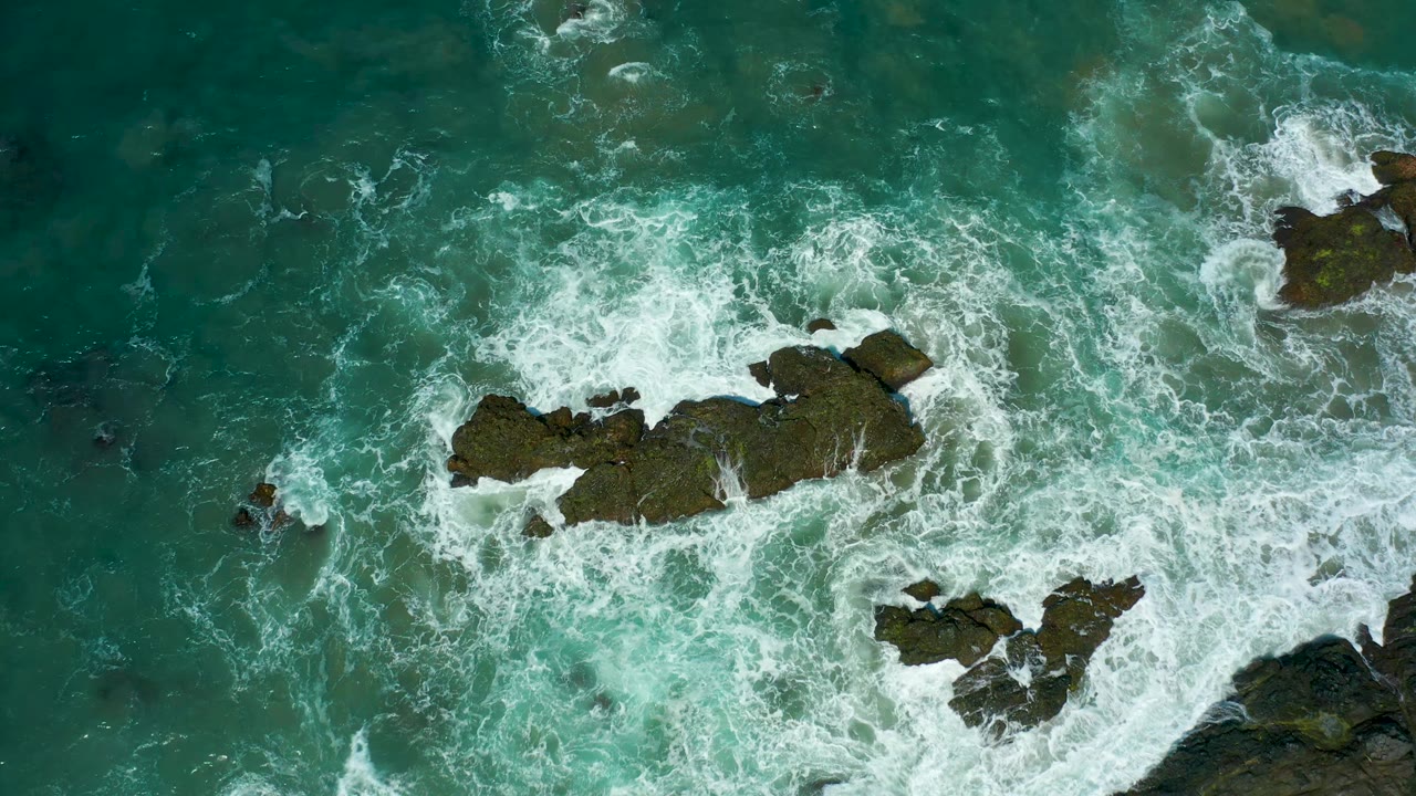 At 0:01, white foam thickens around the central rock formation in the turquoise water at Cerritos Beach, with no visible people or vessels.