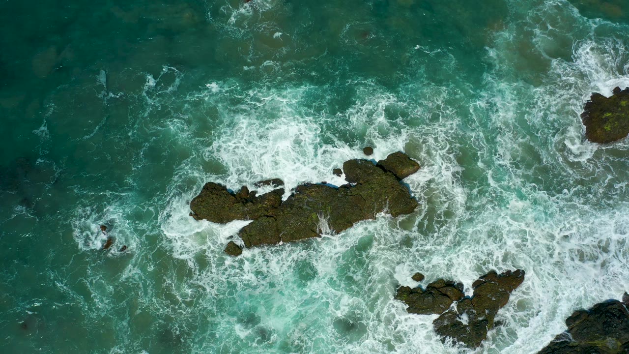 Aerial view of surf curling around rocks in green-blue ocean water.