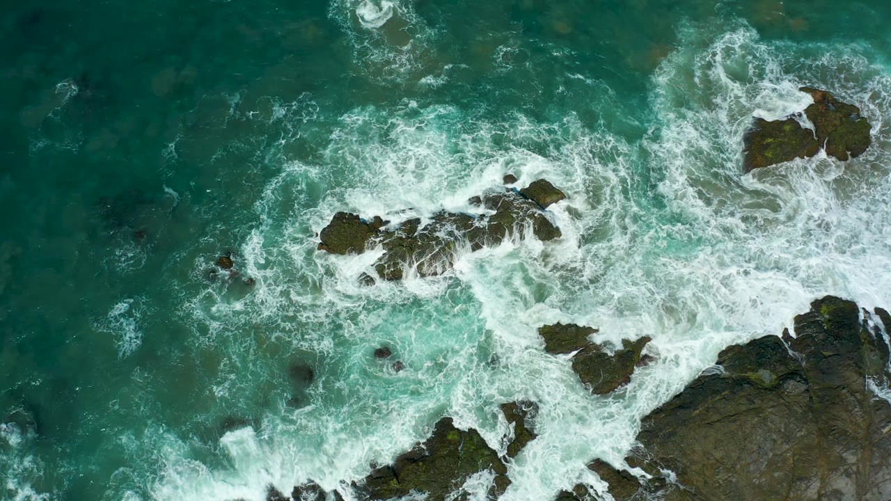 Aerial frame at Cerritos Beach with a broad sheet of white foam crossing the central rocks and turquoise water filling the upper frame, with no visible people.