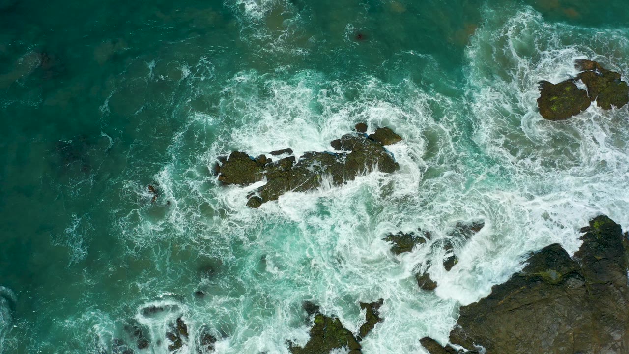 Top-down Cerritos Beach frame with a long dark rock formation near center, foam curling around its sides, and green-blue water surrounding it, with no visible people.