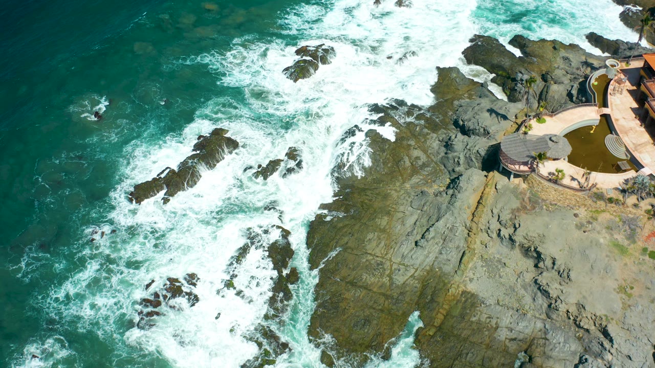 At Cerritos Beach, Baja California Sur, white foam gathers around exposed rock outcrops, with the shoreline pool structure clipped along the right side and no visible people.